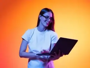 A woman smiling using a laptop with an orange background.